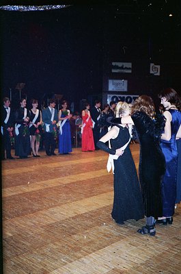 Vintage formal dance event with attendees in 1970s-era attire: men in tuxedos, women in floor-length gowns. Wooden dance floo...