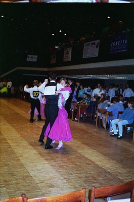 Couple ballroom dancing in a dimly lit hall, likely 1980s–1990s. Man in black suit, woman in pink ballgown. Spectators seated...