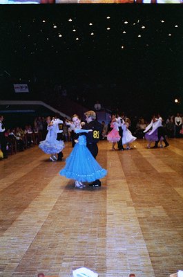 Group ballroom dance competition in a dimly lit hall, featuring children in formal attire. Boys wear suits with numbered bibs...