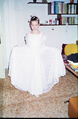 Vintage indoor portrait of a young girl in a voluminous lace wedding dress, posed on a patterned carpet. Bookshelves with col...