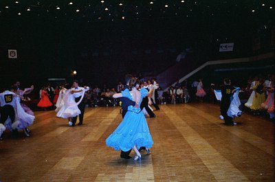 Indoor ballroom dance competition featuring couples in traditional folk attire. Men in dark suits with numbered bibs (, ) pai...