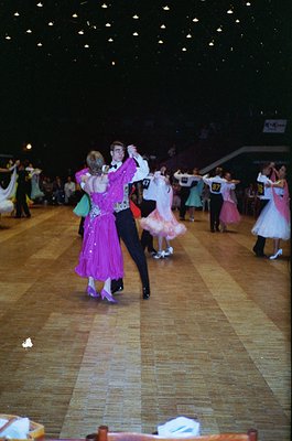Indoor folk dance performance in a grand hall, likely 1980s–1990s Eastern Europe. Couples in traditional attire—women in flow...