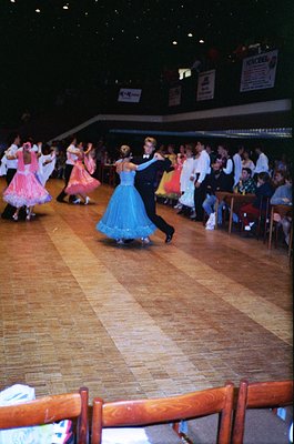 Ballroom dancers in 1970s-style attire perform on polished wooden floor. Couple in blue and black formalwear leads; spectator...