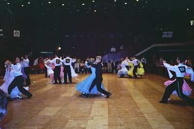 Ballroom dance competition featuring couples in 1970s-style attire—men in white jackets, black pants, and numbered bibs; wome...