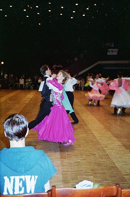 Ballroom dancers in 1990s-style attire perform indoors under bright stage lighting. Couples in coordinated outfits—men in dar...