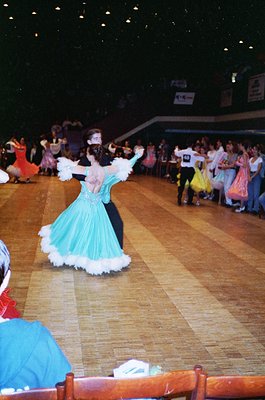 Indoor folk dance performance in a large hall, featuring a woman in a turquoise and black embroidered dress with white ruffle...