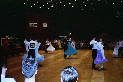 Indoor folk dance performance in a large hall, likely from the 1970s–1980s. Couples in traditional attire—men in white shirts...