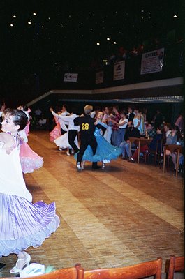 Ballroom dancers in 1970s-style attire perform indoors, likely at a competition or social event. Wooden floor, tiered seating...