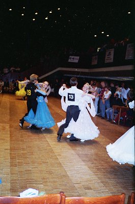 Ballroom dancers in formal attire perform indoors, likely a 1980s-90s competition. Men in black suits with numbered bibs (, )...