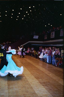 Ballroom dancers in 1970s-style attire perform under string lights in a spacious indoor venue. The woman wears a turquoise go...