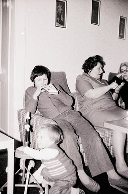 Vintage black-and-white indoor scene featuring three generations: a young boy eating, an adult woman knitting, and a toddler ...