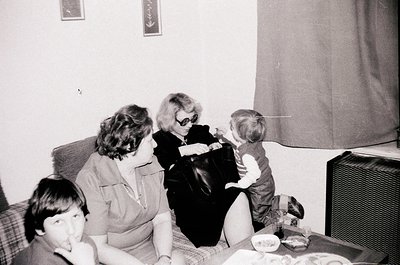 Black-and-white snapshot of an indoor family moment, likely mid-20th century. Four individuals—two adults and two children—ga...