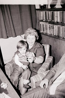 A mid-20th-century indoor portrait showing a woman holding a young child on her lap in a cozy living room. She wears a patter...