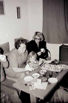 Mid-20th century indoor family gathering at a wooden table with plates of food, including pastries and a cake. Three women an...