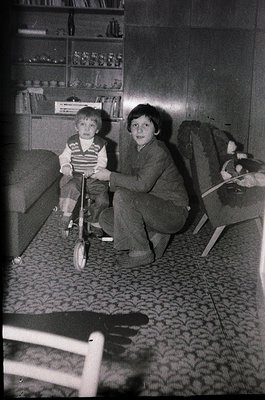Mid-20th century indoor scene: two children in a dimly lit living room, one seated on a balance bike, the other kneeling besi...