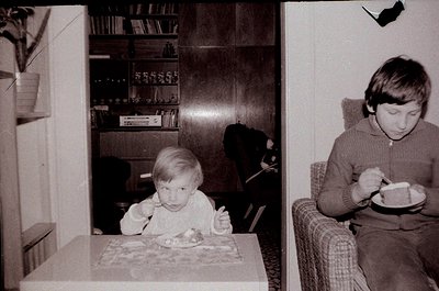 Two children eating indoors, likely mid-1970s. Child on left sits at a patterned tablecloth-covered table with a spoon, surro...