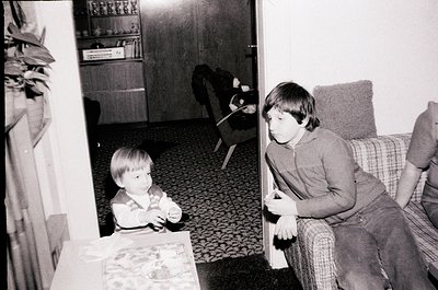 Two children in a mid-century living room, one seated on patterned sofa, the other standing by a low table with a patterned c...