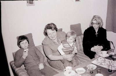 Black-and-white snapshot of a mid-20th century family gathering, likely 1960s–1970s. Three adults and a child seated on a pat...