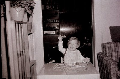Vintage black-and-white photo of a child (approx. 2-3 years) seated at a small table, holding a spoon with cake. Mid-century ...
