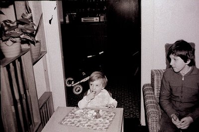 Vintage black-and-white photo of two children in a mid-century living room. Toddler seated at a round table with a floral-pat...