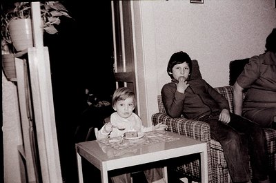 Two children seated indoors at a small table, likely mid-20th century. Infant in striped shirt holds a cake with lit candles;...