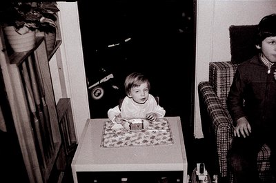 A toddler sits at a small wooden table in a mid-century interior, wearing a white long-sleeve shirt and patterned pants. Behi...