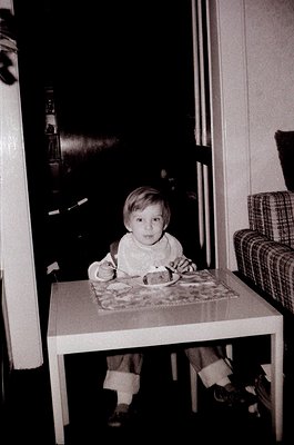 Young child seated at a small wooden table in a mid-century kitchen, holding a plate with cake. Checkered tablecloth and plai...