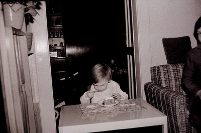Vintage black-and-white photo of a toddler seated at a highchair, eating from a paper plate with a cartoon character. Mid-cen...