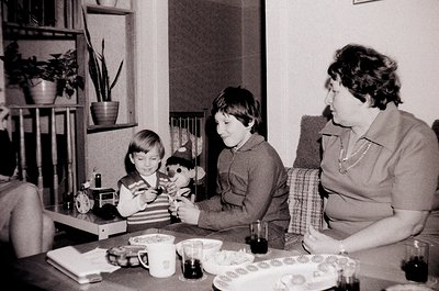 Three children and an adult share a mid-century indoor moment, likely 1960s–1970s. A boy in a sweater plays with a stuffed an...