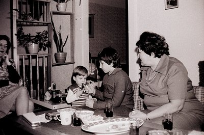 Vintage black-and-white indoor scene featuring a woman in a collared blouse and two boys at a table, likely mid-1970s. The wo...