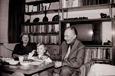 A mid-20th-century indoor family moment: elderly couple and child seated at a small wooden table, surrounded by bookshelves f...