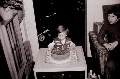 Vintage black-and-white indoor scene: young child seated at a small table blowing out a single candle on a decorated sheet ca...