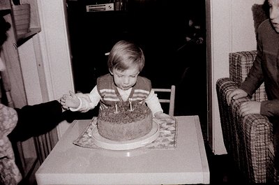 Young child blowing out candles on a round chocolate cake, seated at a small table in a mid-century indoor setting. Patterned...