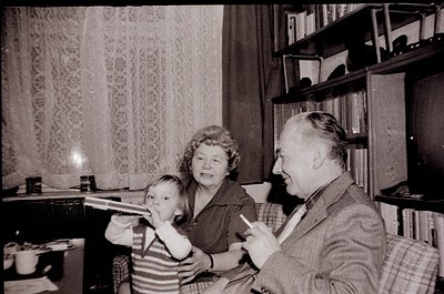 Mid-20th century (1950s–60s) indoor family moment: elderly couple and child in a cozy living room with patterned curtains, wo...