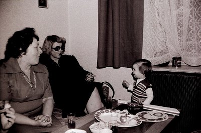 Three generations share a meal in a mid-century indoor setting—two women in formal attire (one with sunglasses indoors) and a...