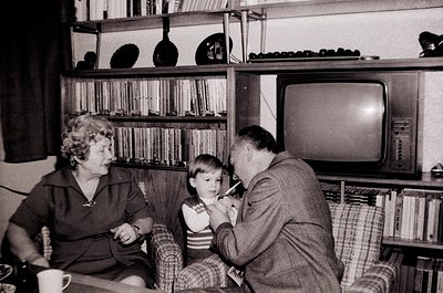 Vintage black-and-white interior shot of a mid-century living room. A man in a suit assists a young child with brushing teeth...