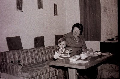Vintage indoor scene featuring a woman and young boy seated at a small wooden table in a modest living room. Plaid sofa and f...