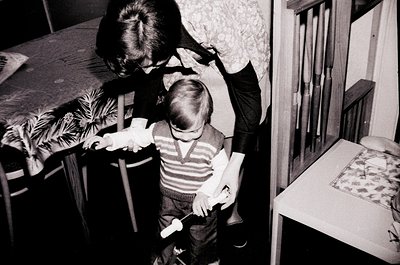 Black-and-white interior shot of a child (front) and adult (back) in a modest home, likely mid-20th century. The child stands...
