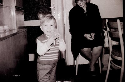 Mid-century kitchen scene: boy in striped sweater and shorts poses with toy, woman in skirt and glasses sits on stool. Wooden...
