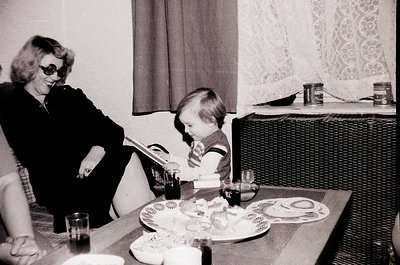 A mid-20th-century indoor scene captures a woman in sunglasses reading beside a child seated at a table. The table holds vint...