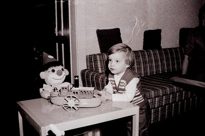 Vintage black-and-white photo of a young child seated at a small table, playing with a toy train set featuring a smiling char...