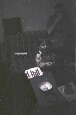 Vintage black-and-white photo of a child playing with a toy train set on a striped sofa. Reflections and scratches obscure cl...