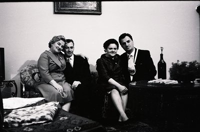 Four individuals pose indoors in formal attire, likely 1950s–1960s. Men wear suits with ties; women in structured dresses. Bo...