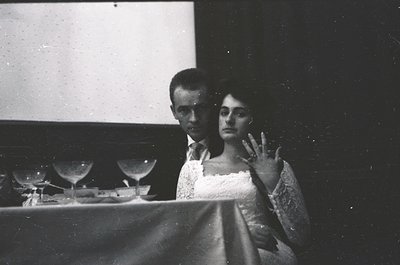Couple seated at a table in a dimly lit, speckled projection room, likely a 1950s–1960s cinema. Man wears a dark suit; woman ...