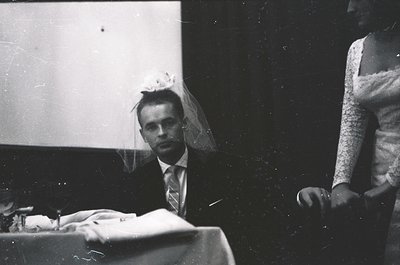 Black-and-white portrait of a groom in a tuxedo with veil, seated at a table with a white cloth. Partial view of bride in lac...