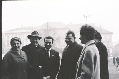 Five individuals pose outdoors in 1960s Eastern Bloc attire—men in suits with badges, women in coats and hats—against a Sovie...