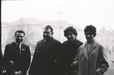 Four individuals pose outdoors in mid-20th century attire, likely 1950s–1960s. The man on the left wears a pilot’s jacket wit...