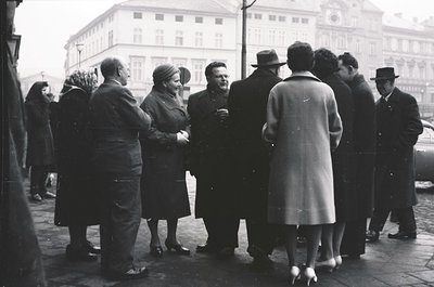 Group of 12+ men and women in formal 1940s attire—overcoats, hats, gloves—standing outdoors near a grand building with classi...