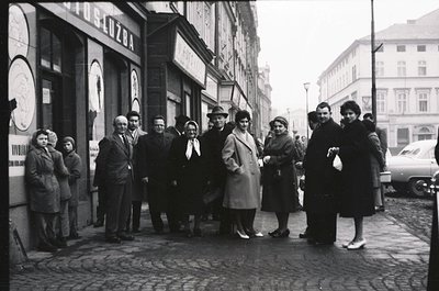 Vintage group portrait on a European city street, likely 1950s–1960s. Nine individuals in mid-century attire—coats, hats, and...