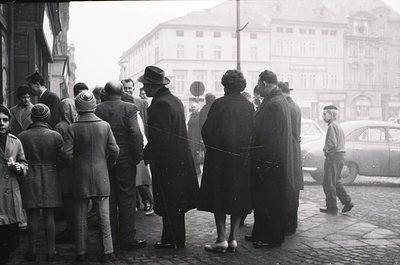 A mid-20th century street scene featuring a diverse crowd in winter attire, lined up outside a building with ornate facades. ...